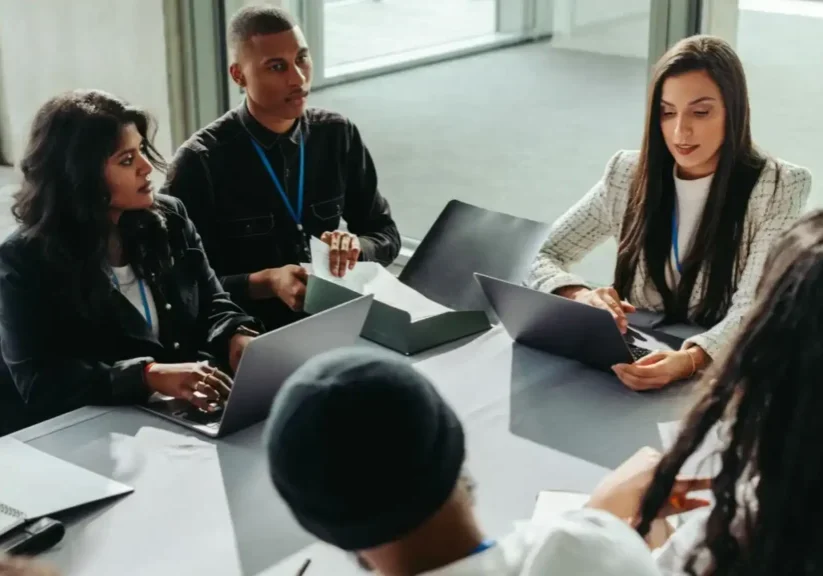A diverse group discussing work around a table with laptops.